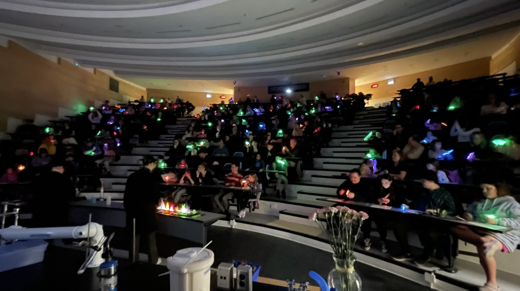 Image of seated audience with glowsticks while student performs flaming bowls chemistry experiment